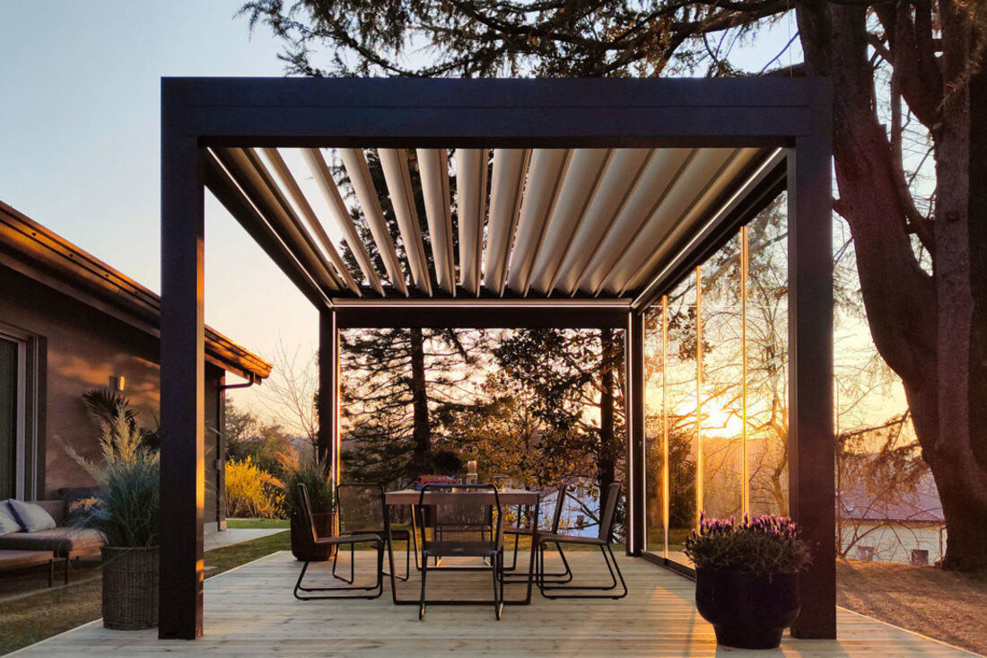 Pergola bioclimatica antracite con lame aperte, al tramonto, sopra un tavolo da pranzo esterno